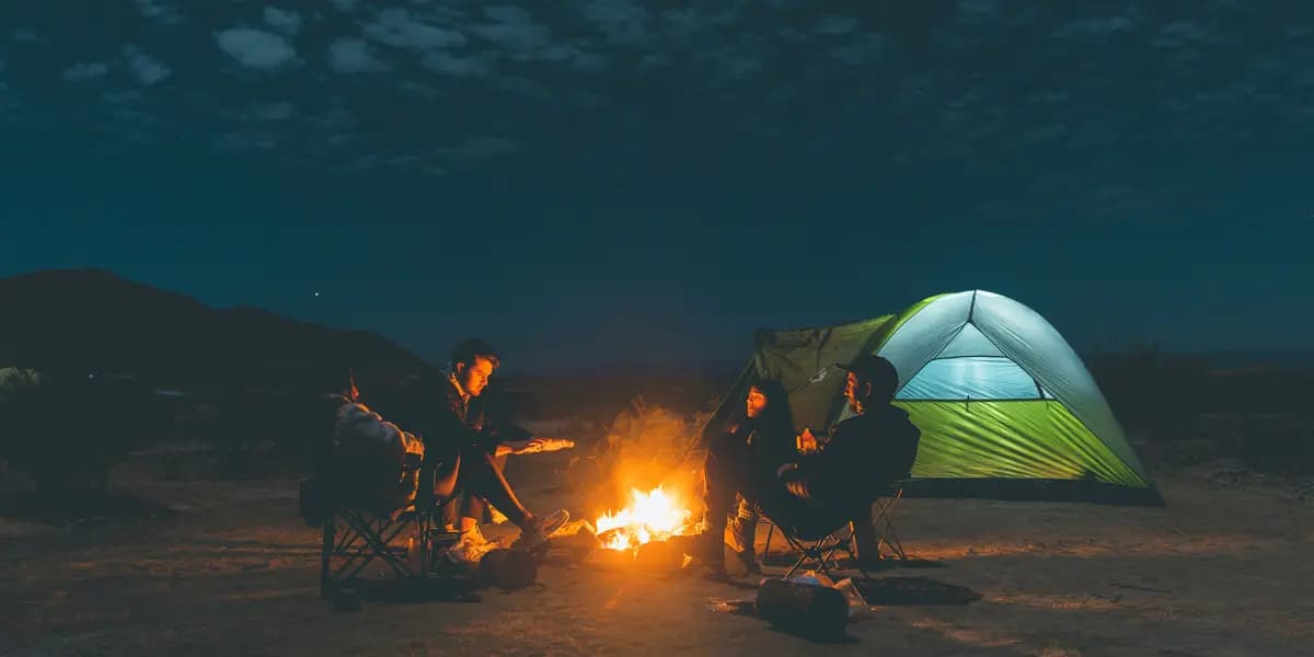 Παρέα σε camping τη νύχτα γύρω από φωτιά δίπλα σε φωτισμένη σκηνή